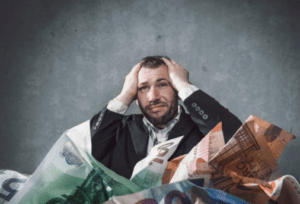 Stressed man in suit holding head surrounded by piles of oversized foreign currency